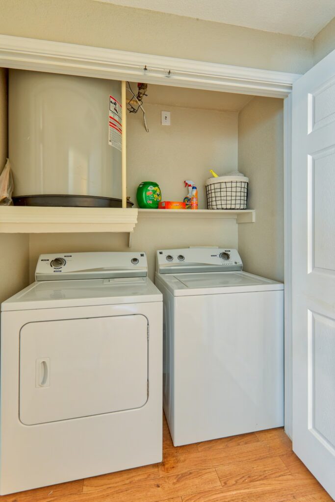 A laundry closet with a white washer and dryer, shelf above holding detergent, cleaning supplies, and a basket, and a water heater in the upper left corner.