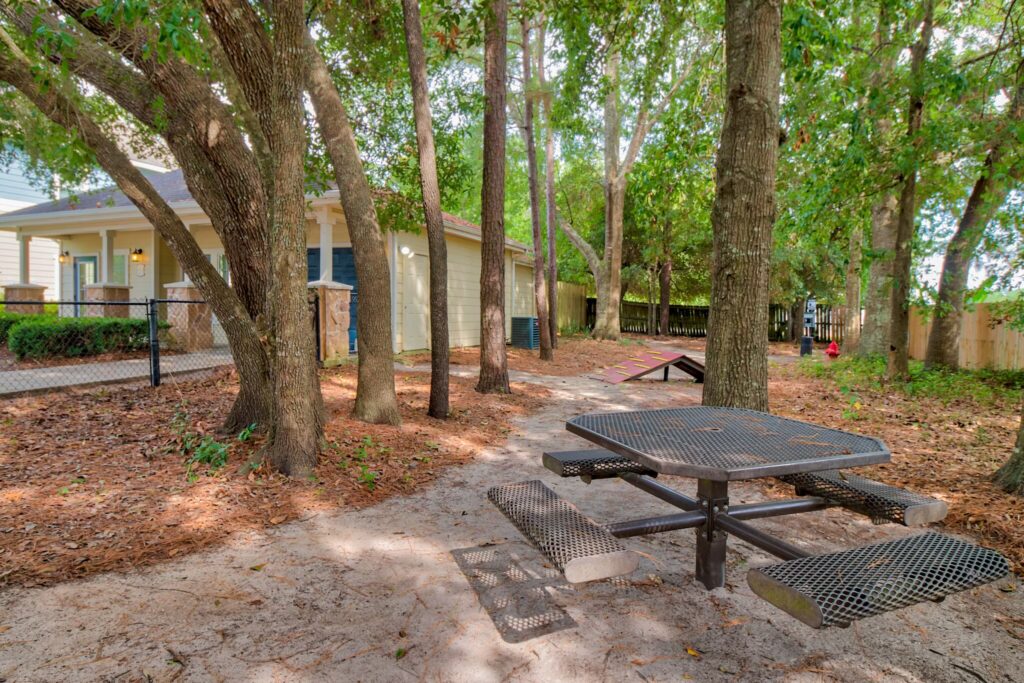 A metal picnic table sits under several trees on a dirt surface near a fenced yellow building in a shaded outdoor area.