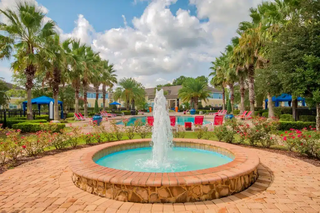 A round stone fountain is centered in a landscaped area with blooming bushes, palm trees, red lounge chairs, and a swimming pool in the background under a partly cloudy sky.